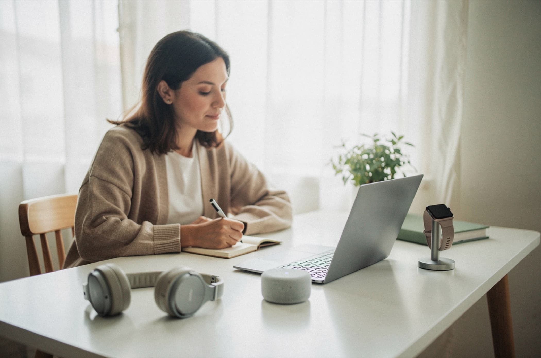 A reviewer at her desk writing about the headphones, speaker and smartwatch laid out in front of her